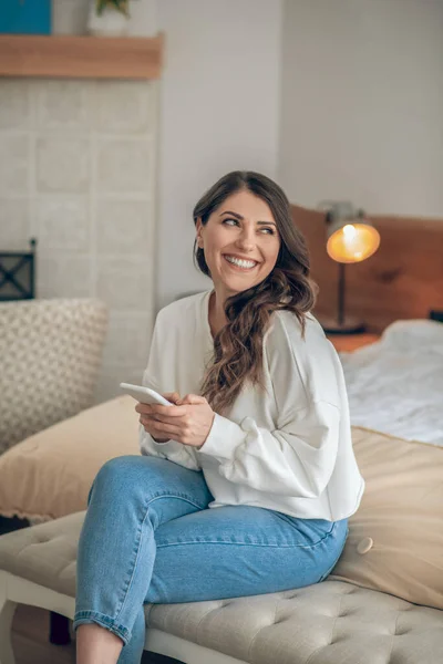 Giovane donna sorridente con uno smartphone in mano seduta sul letto — Foto Stock