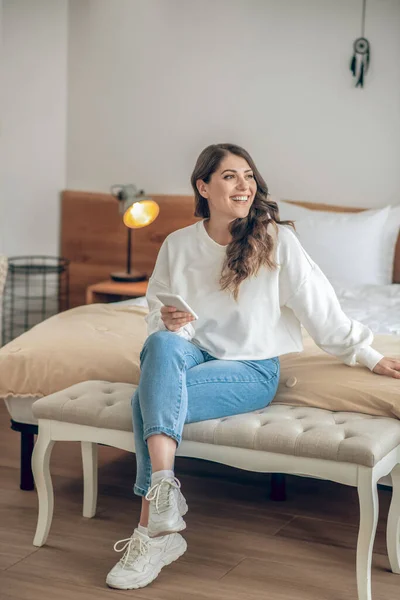 Jovem sorridente com um smartphone nas mãos sentado na cama — Fotografia de Stock