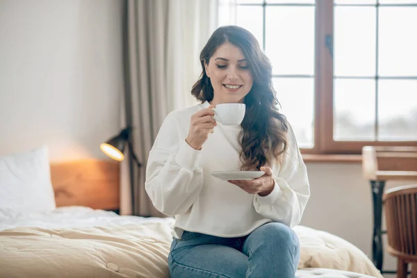 Wanita muda cantik duduk di tempat tidur dan minum kopi — Stok Foto