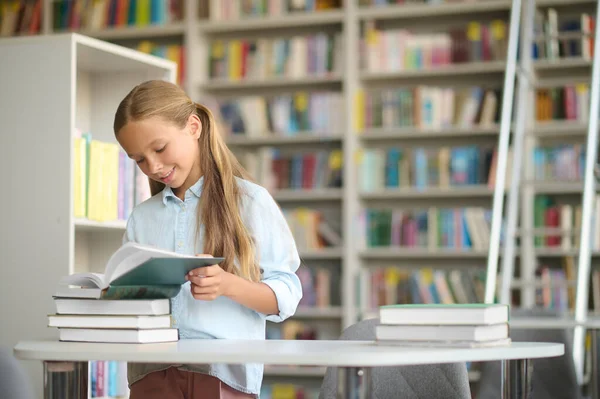 Glad elev läser en lärobok vid bibliotekets skrivbord — Stockfoto