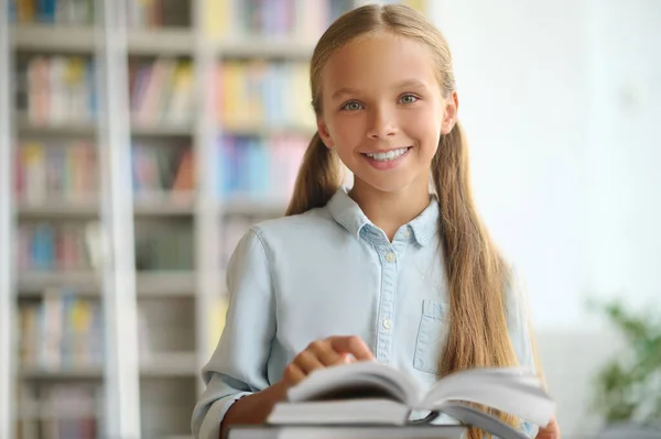 Kitaplarla dolu bir masada duran mutlu şirin okul çocuğu. — Stok fotoğraf