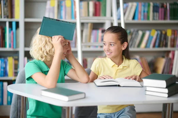 Söt flicka och hennes glada vän sitter på ett bibliotek — Stockfoto