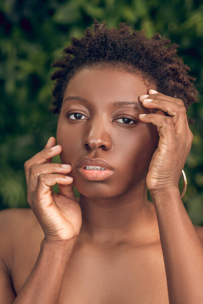 Skin care. Young adult attractive african american woman with bare shoulders touching face against green plants background