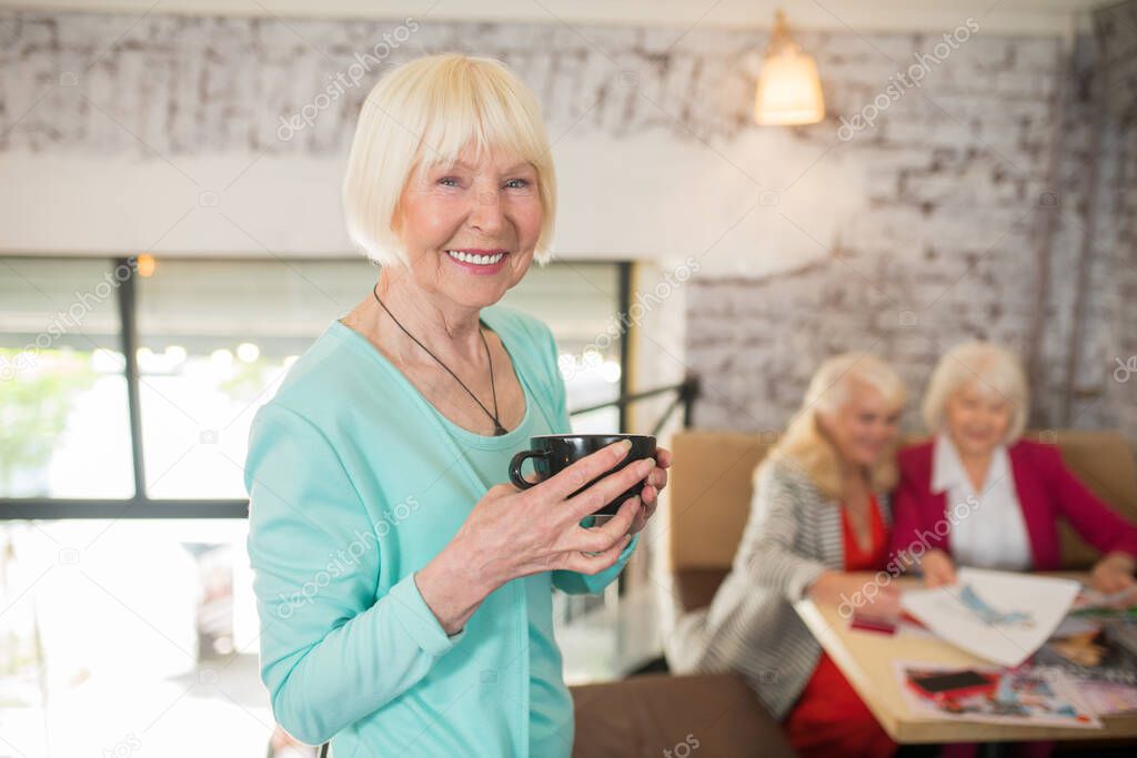 A blonde woman in a blue suit standing with a cup of tea in hands and smiling