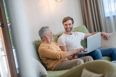 Genç sakallı bir adam dizüstü bilgisayarda çalışıyor, babası yanında oturuyor.