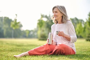 Termoslu bir kadın parkta çimlerde oturuyor.
