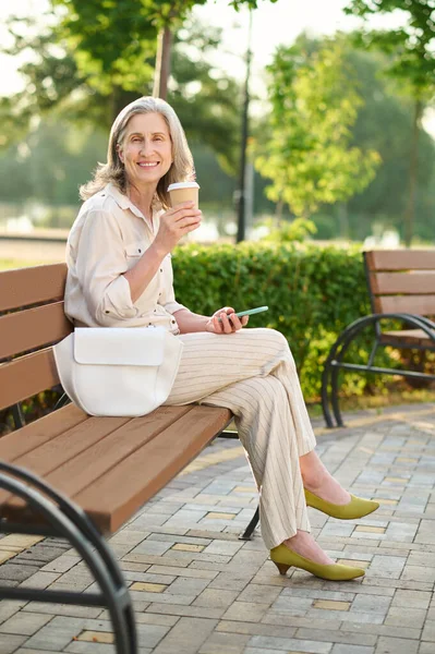 Добре виглядає жінка п'є каву на лавці парку — стокове фото