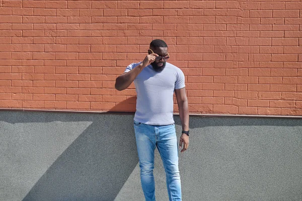 Un hombre de piel oscura con una camiseta blanca y jeans cerca de la pared de ladrillo — Foto de Stock