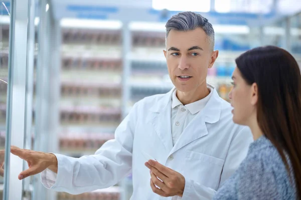 Frau und aufmerksamer Apotheker vor Regalen mit Medikamenten — Stockfoto