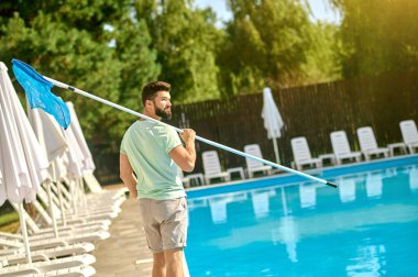 Koyu saçlı bir adam yüzme havuzunu temizliyor ve meşgul görünüyor.