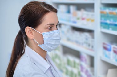 Koruyucu maskeli ve beyaz önlüklü düşünceli bir kadın.