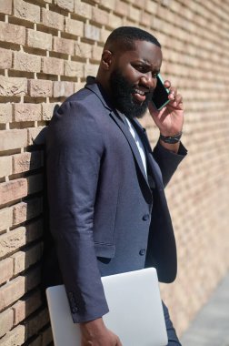 Tuğla duvarın yanında takım elbiseli Afro-Amerikan bir adam duruyor.