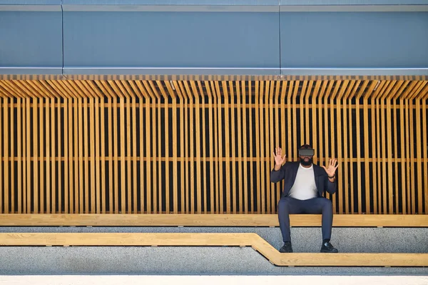 VR gözlüklü koyu tenli bir adam bankta oturuyor.