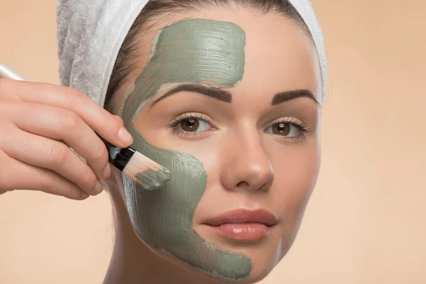 Spa girl with a  towel on her head applying facial clay mask and — Stock Photo, Image