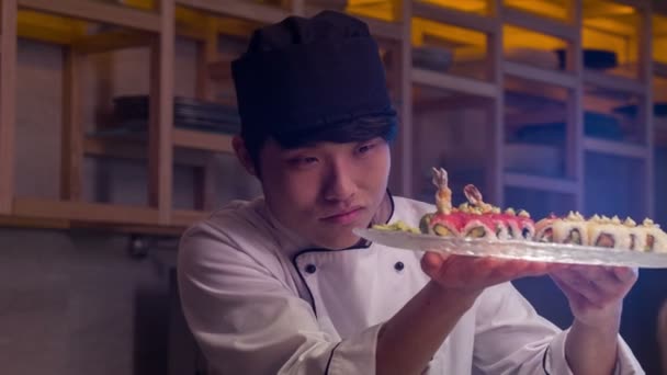 Sorrindo chef asiático em uniforme branco de pé na cozinha de sushi bar transformando placa com rolos — Vídeo de Stock