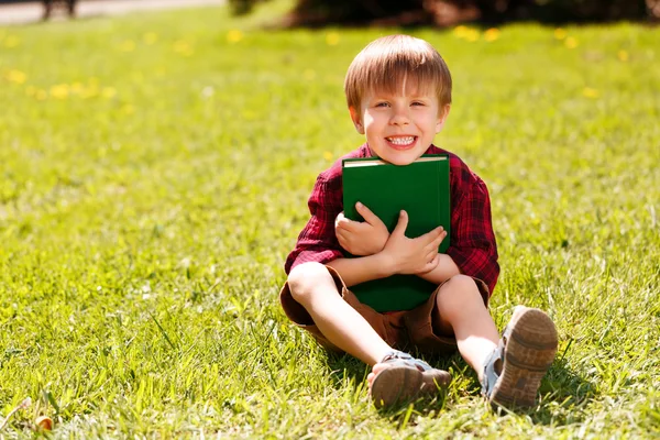 Sonriente niño sentado en la hierba y abrazando libro — Foto de Stock