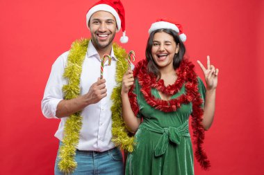 Noel Baba şapkası takıp yeni yılı birlikte kutlayan mutlu çift kışın kırmızı arka planda saklanmış şeker kamışları tutuyor.