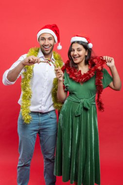 Noel Baba şapkası takıp yeni yılı birlikte kutlayan mutlu çift kışın kırmızı arka planda saklanmış şeker kamışları tutuyor.