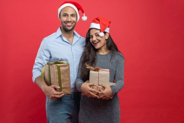 Afro-Amerikalı erkek ve Hintli kadın, yeni yılı kırmızı arka planda izole edilmiş hediye kutuları ve süslemelerle kutluyorlar.