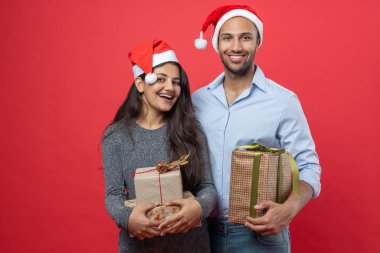 Mutlu çift, Noel kutlamaları sırasında kırmızı arka planda izole edilmiş şenlikli şapkalar ve süslemelerle hediye kutuları taşıyor.