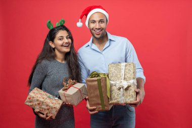 Noel arifesini kutlayan hediye kutuları ve Noel süsleri taşıyan aile, kışın kırmızı arka planda izole edilmiş şenlikli şapkalar takıyor.