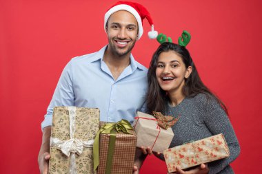 Mutlu çift, Noel kutlamaları sırasında kırmızı arka planda izole edilmiş şenlikli şapkalar ve süslemelerle hediye kutuları taşıyor.