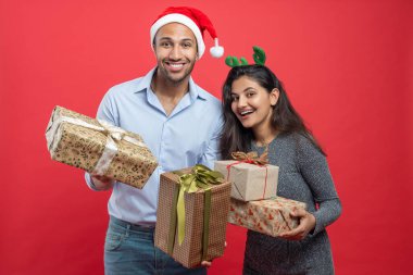 Mutlu çift, Noel kutlamaları sırasında kırmızı arka planda izole edilmiş şenlikli şapkalar ve süslemelerle hediye kutuları taşıyor.
