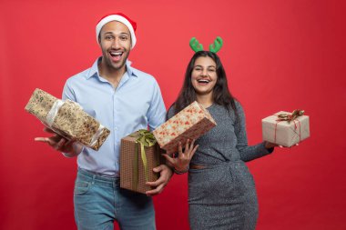 Noel arifesini kutlayan hediye kutuları ve Noel süsleri taşıyan aile, kışın kırmızı arka planda izole edilmiş şenlikli şapkalar takıyor.