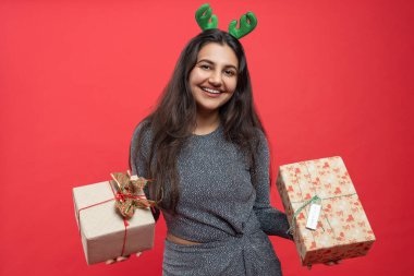 Ren geyiği boynuzu takmış bir kadın Noel partisinde kırmızı arka planda izole edilmiş hediye kutularını tutarak mutlu noeller kutluyor.