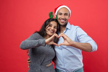Mutlu çift, kırmızı arka planda izole edilmiş Noel kutlaması sırasında kalp şeklini birlikte gösteriyor.
