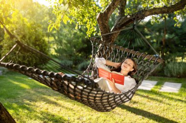 Boş zaman. Uzun saçlı, beyaz, genç bir kadın hamakta elinde kitapla keyif çatıyor.