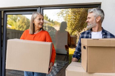 Sundurmada kutuları birlikte taşıyan yaşlı çift gülümsüyor ve taşınma gününün tadını çıkarıyor.