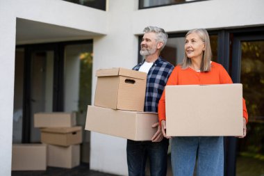 Taşınma günü boyunca evin önünde gülümseyen kutuları taşıyan olgun bir çift.