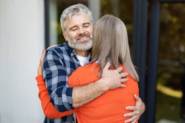 Sarılalım. Gri saçlı sakallı adam karısına sarılıyor ve mutlu bir şekilde gülümsüyor.