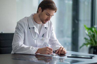 Genç doktor açık pencerenin yanında tıbbi kayıtlar yapmaya odaklanmış.