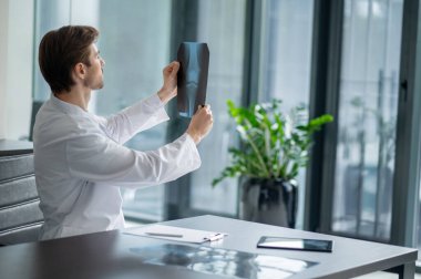 Masada oturan erkek doktor geniş pencereli modern klinikteki röntgenlere bakıyor.