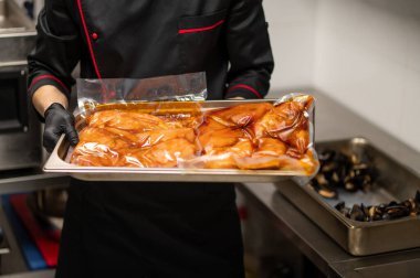 Siyah üniformalı tanınmayan bir aşçı mutfakta metal tepsi ve marine edilmiş yemek tutuyor.