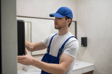 Erkek bakım teknisyeni fayanslı duvara yeni el kurutma makinesi yerleştiriyor.