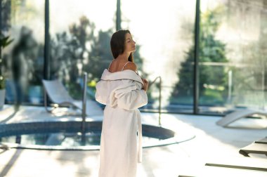 Rahatladım ve eğlendim. Beyaz banyo bornozu giymiş bir kadın havuz kenarında bir spa merkezinde duruyor.