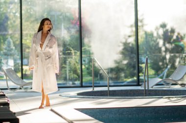 Spa salonunda. Kaplıca salonundaki yüzme havuzunun kenarında uzun bir bornoz giymiş genç bir kadın.