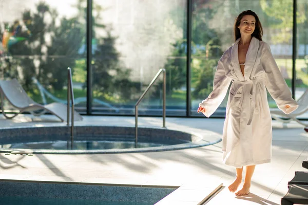 Spa salonunda. Kaplıca salonundaki yüzme havuzunun kenarında uzun bir bornoz giymiş genç bir kadın.