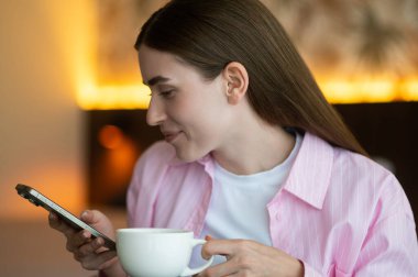 Huzurlu bir sabah. Elinde telefonla çay içen pembe elbiseli hoş bir genç bayan.