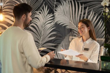 Resepsiyonda. Resepsiyonist erkek bir müşteriyle konuşuyor ve ona kalacak yer ayarlıyor.