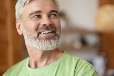 Gri saçlı, sakallı, mutfakta gülümseyen yetişkin bir adam.