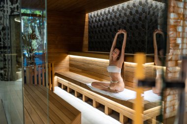 Sauna zamanı. Slim fit genç kadın saunada bankta uzanıyor ve rahatlamış görünüyor.