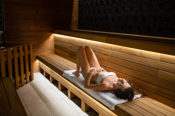 Sauna zamanı. Slim fit genç kadın saunada bankta uzanıyor ve rahatlamış görünüyor.