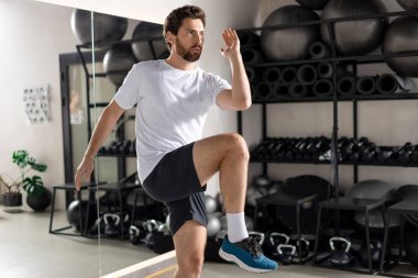 Spor salonunda. Sakallı genç adam spor salonunda egzersiz yapıyor ve odaklanmış görünüyor.