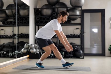 Spor salonunda. Sakallı genç adam spor salonunda egzersiz yapıyor ve odaklanmış görünüyor.