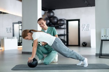 Çekirdek gücü. Küçük kız denge topunda kadın terapist desteğiyle kalas kullanıyor.