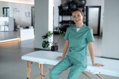 Fizyoterapi seansı. Kanepede oturan kadın fizyoterapist kameraya gülümsüyor ve seansa hazırlanıyor.